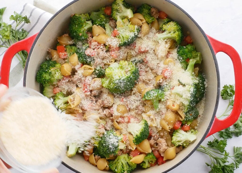 Cheese being poured into pot with broccoli, ground turkey, and small shell pasta.