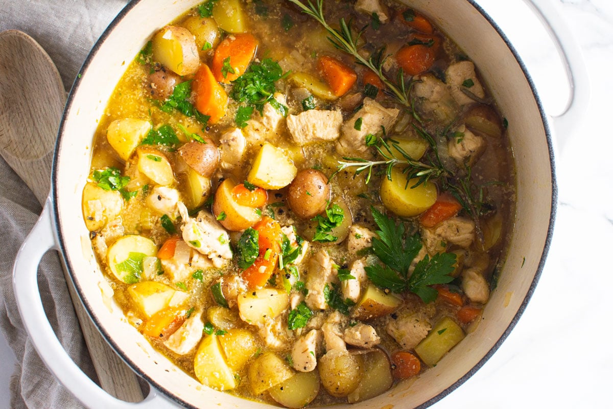 Chicken stew with potatoes, carrots, parsley and rosemary garnish in white pot.