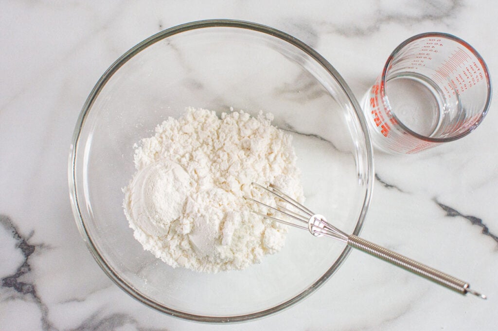 Icing sugar in bowl with whisk and glass of water in measuring cup.