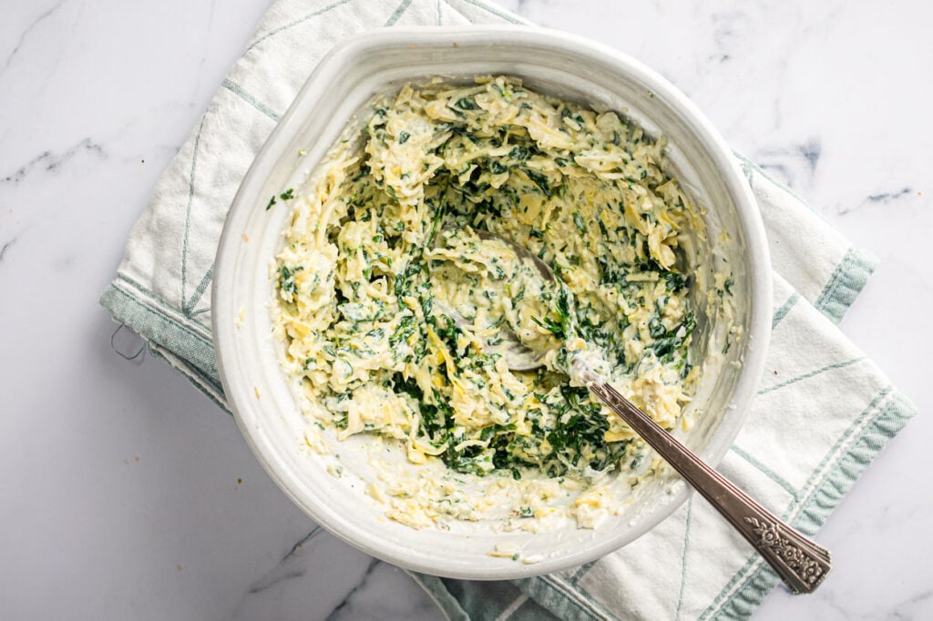 Stirring ingredients for spinach and artichoke dip in white bowl.