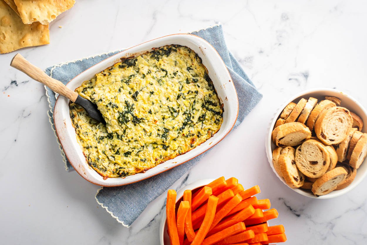 Healthy spinach artichoke dip in baking dish with veggies and crostini in bowls.