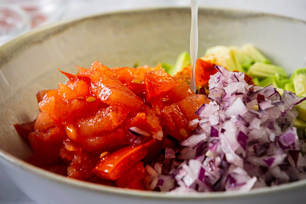 Red onion, chopped tomatoes, avocado in a bowl with lime juice being poured over it.
