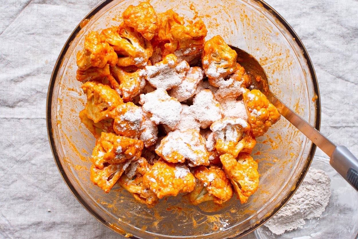 Cornstarch and seasonings sprinkled over buffalo sauce coated cauliflower in a bowl.