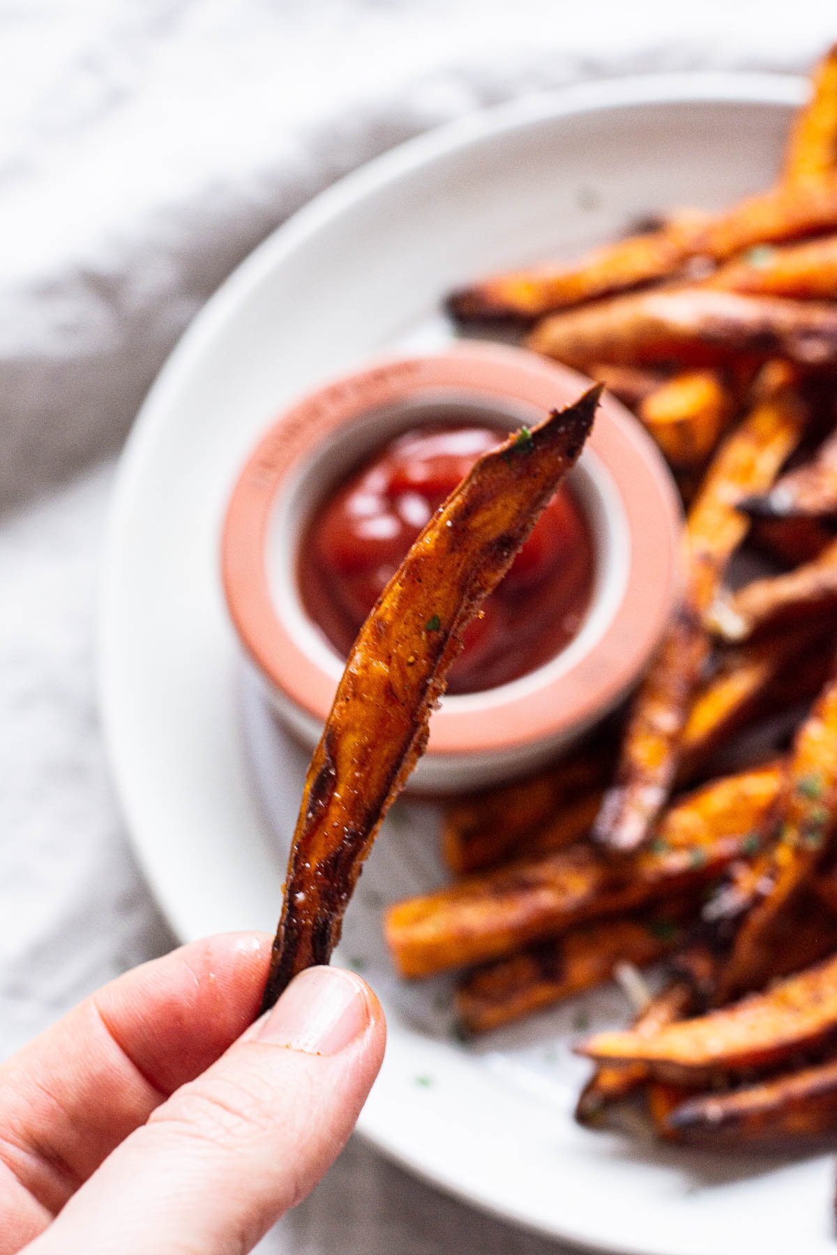 Holding one air fryer sweet potato fry with plated fries in background with ketchup.