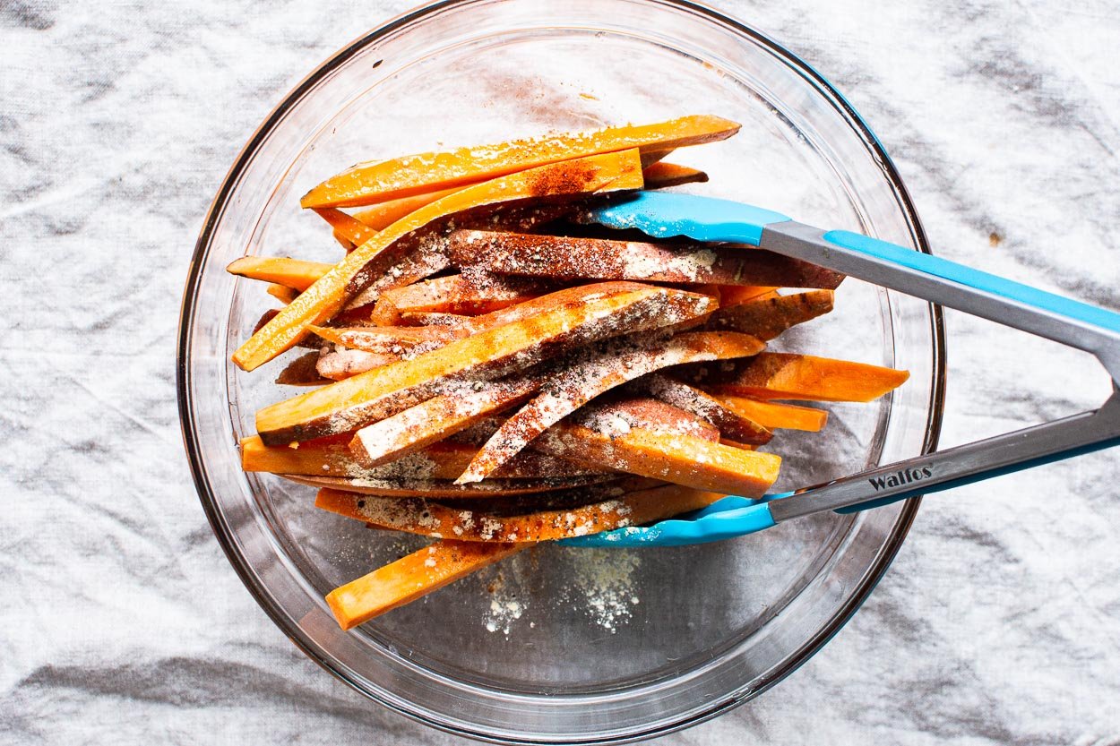 Raw sweet potato fries in a bowl with tongs and seasonings.