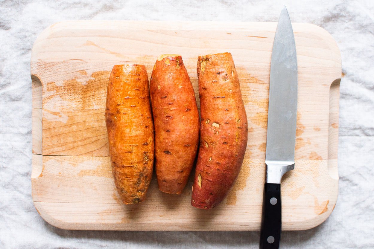 Three sweet potatoes on cutting board with ends sliced off.