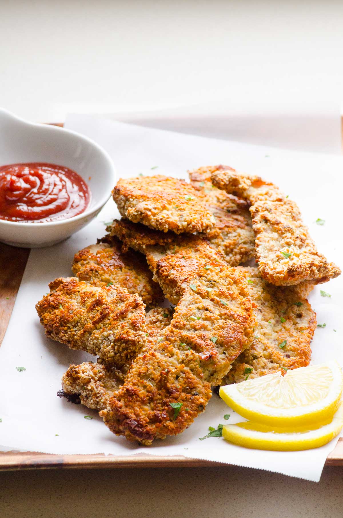 Almond crusted chicken tenders on parchment paper with a lemon wedge and ketchup.