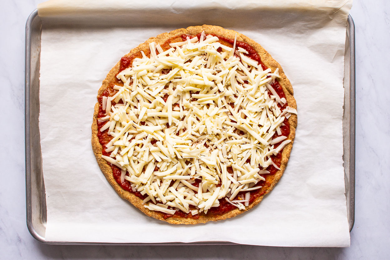 Almond flour pizza crust topped with sauce and cheese on baking sheet.