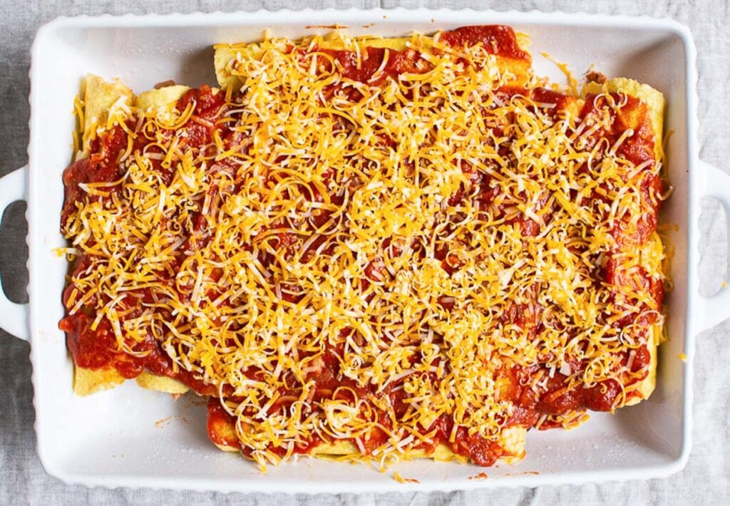Unbaked enchiladas covered with sauce and cheese in a baking dish.