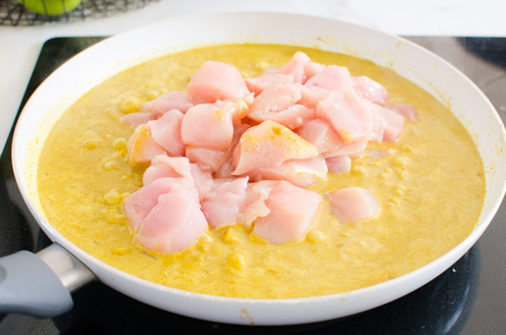 Raw chicken breast pieces added to yellow curry in skillet.