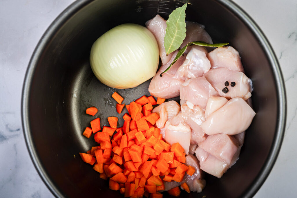 instant pot chicken soup ingredients in inner pot onion, carrots and chicken with peppercorn and bay leaves