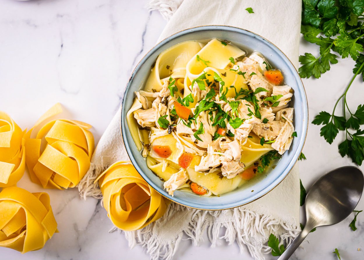 A bowl with instant pot chicken noodle soup and noodles, parsley, and spoon nearby