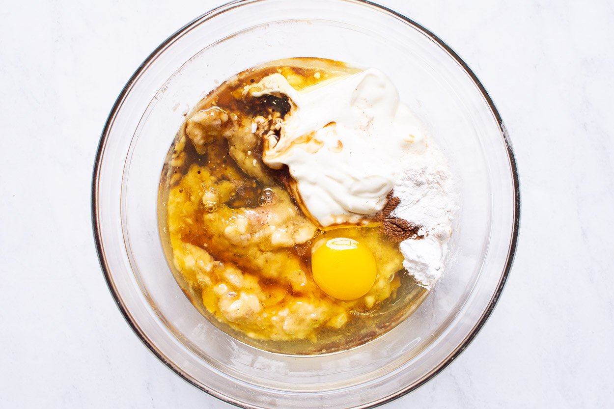 Egg, maple syrup, mashed bananas, oil, yogurt and liquid baking essentials in glass bowl.