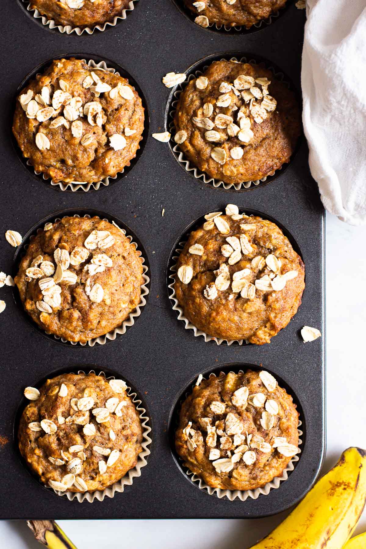Healthy banana oatmeal muffins in muffin tin with a banana and white towel on a counter.