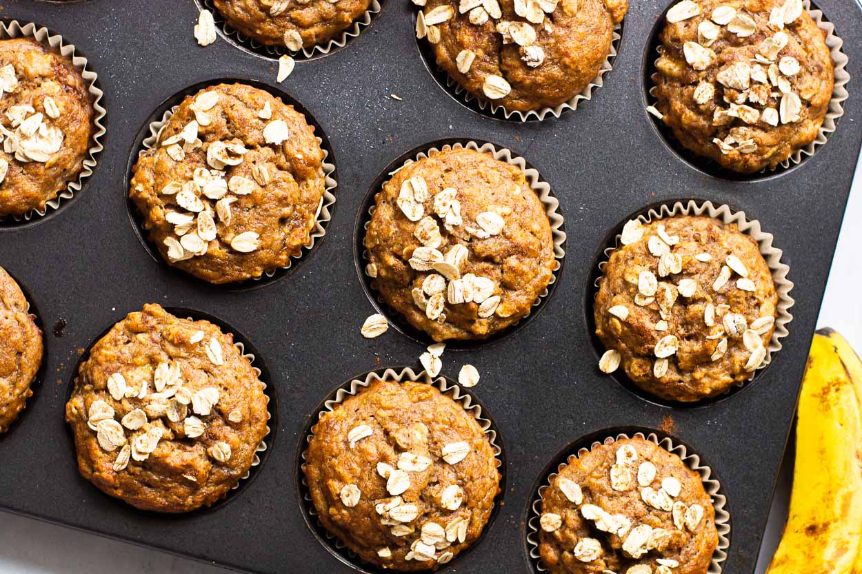 Healthy banana oatmeal muffins in muffin tin with yellow banana beside tin.