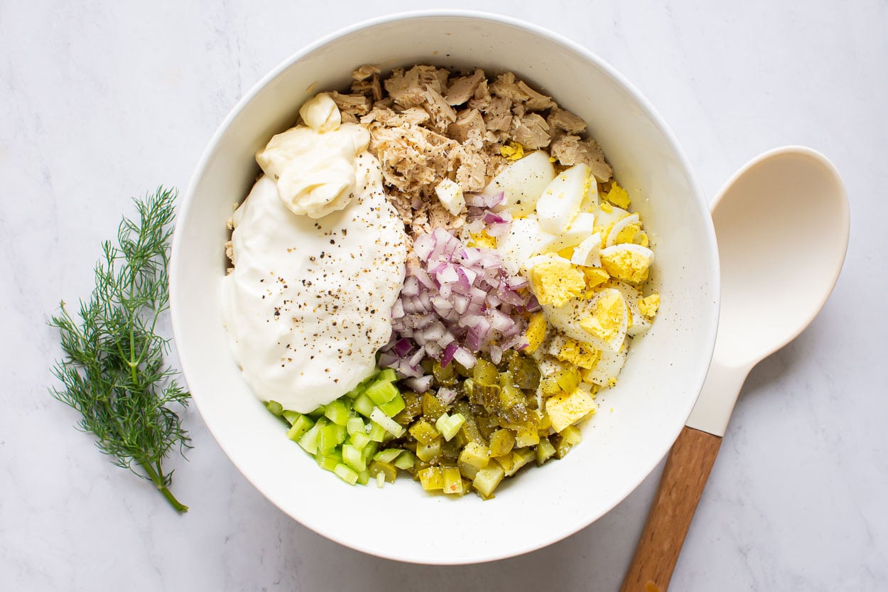 Flaked tuna, chopped egg, celery, pickles, red onion and mayo in white bowl with spoon.