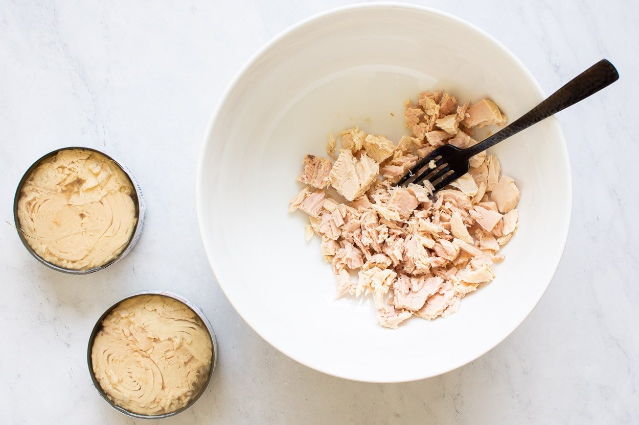 Two cans with tuna and flaked with fork tuna in white bowl.