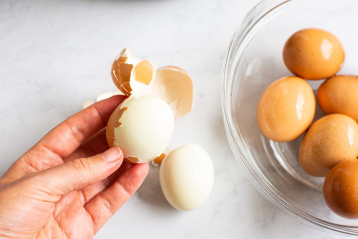 Peeled hard boiled egg with bowl of brown eggs in background.