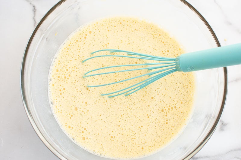 A blue whisk in a bowl of eggs and baking staples that have been mixed.