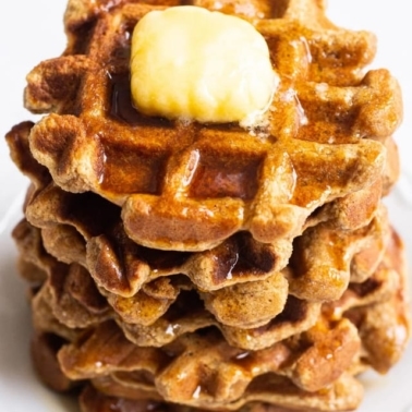 A stack of almond flour waffles on a white plate with butter and maple syrup on top.