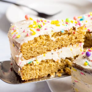 A slice of healthy birthday cake layered with icing and sprinkles being served with a plate in background.