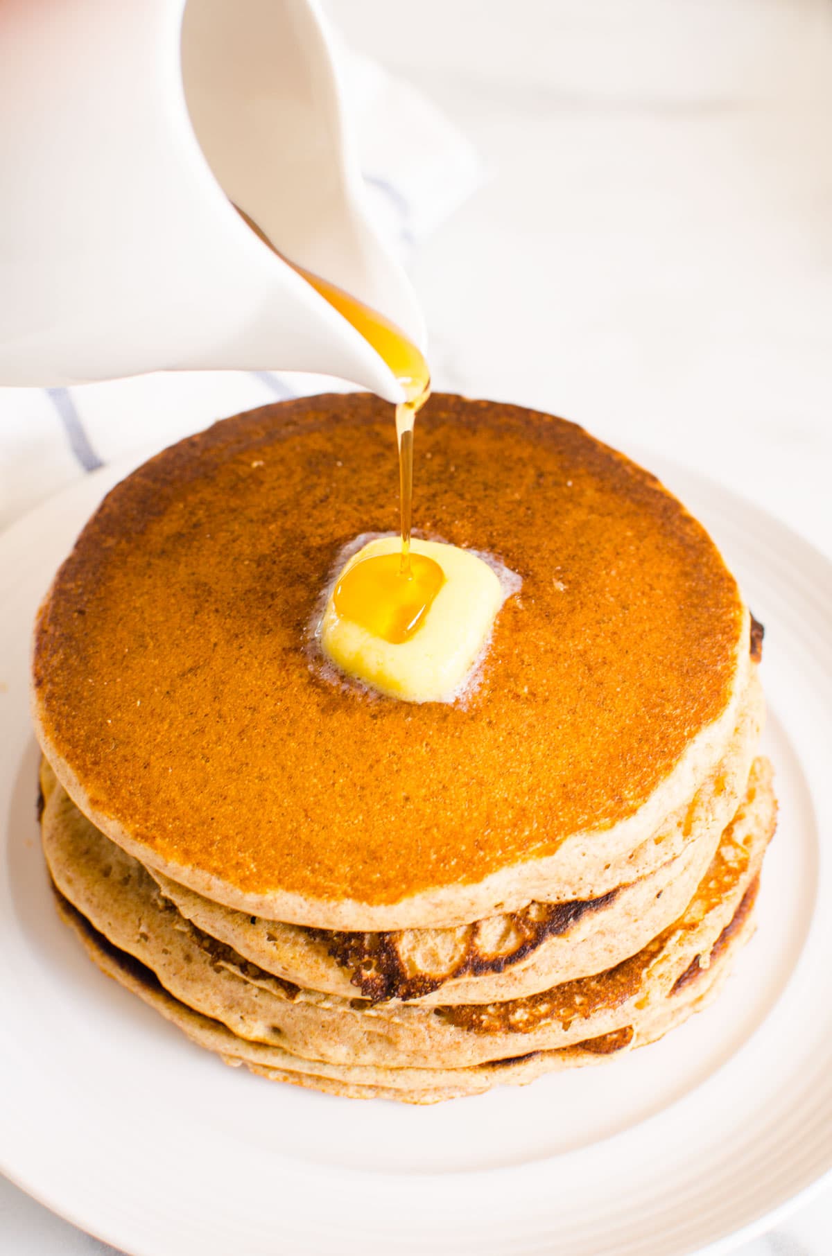 Pouring maple syrup over a stack of whole wheat buttermilk pancakes with butter.