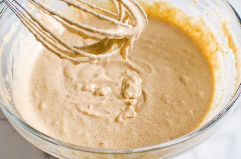 Whole wheat pancake batter dripping from a whisk over a glass bowl.