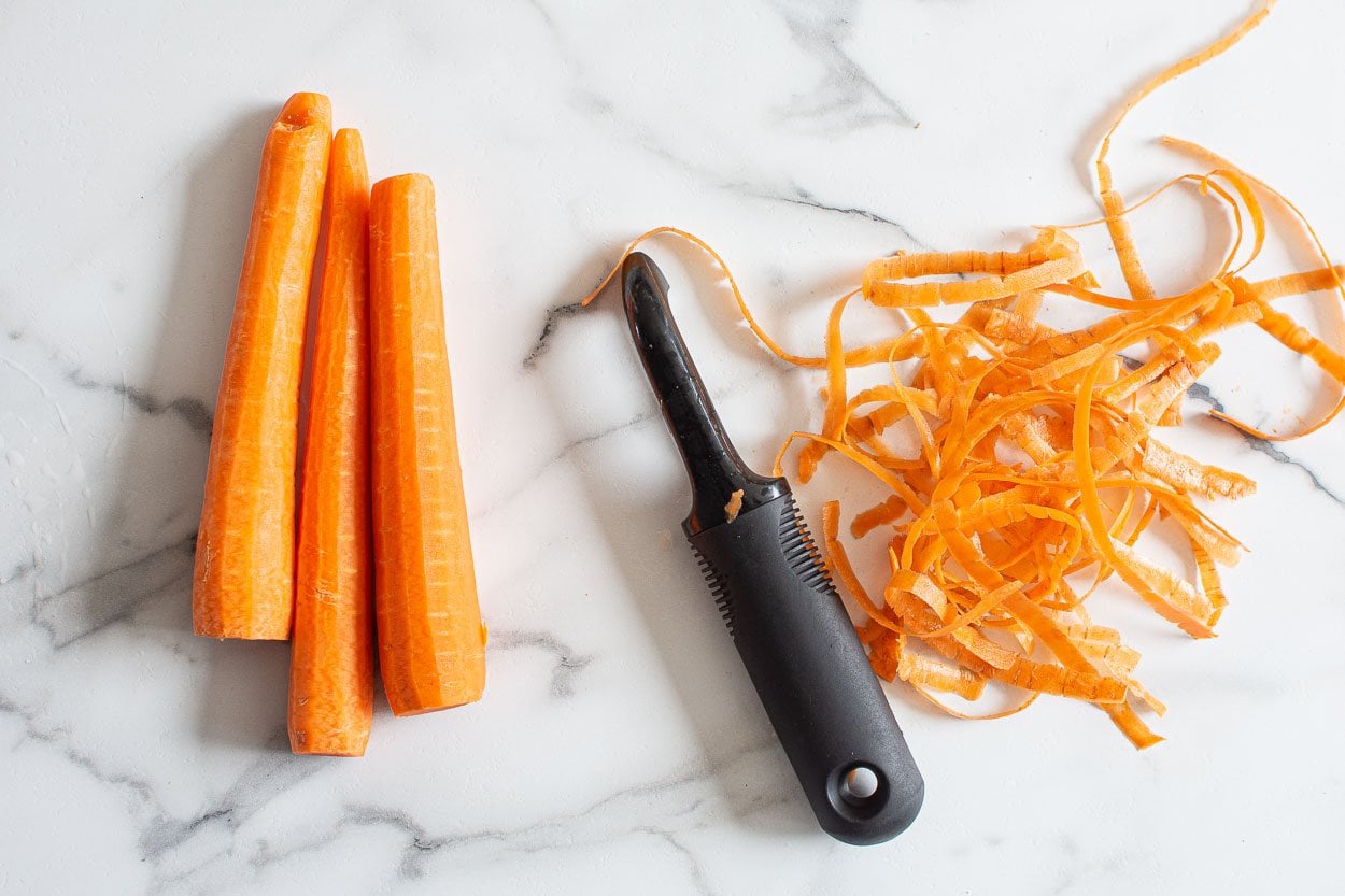 Carrots with vegetable peeler and peels on a counter.