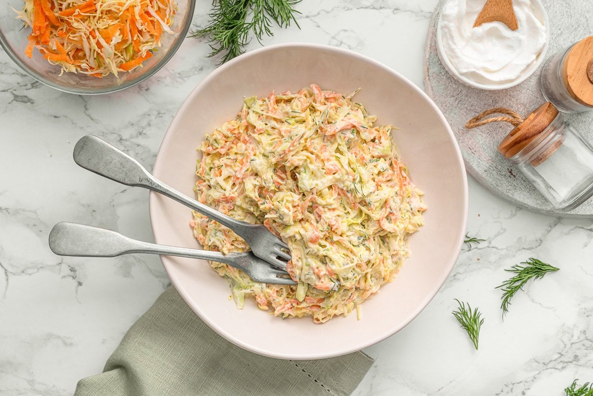 Healthy coleslaw in a bowl with 2 forks. Ingredients on a countertop.
