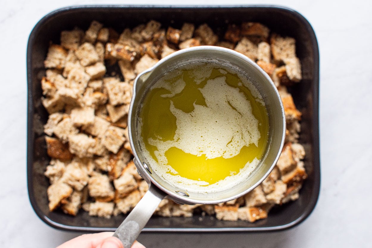 Pouring melted butter over bread cubes in a baking dish.