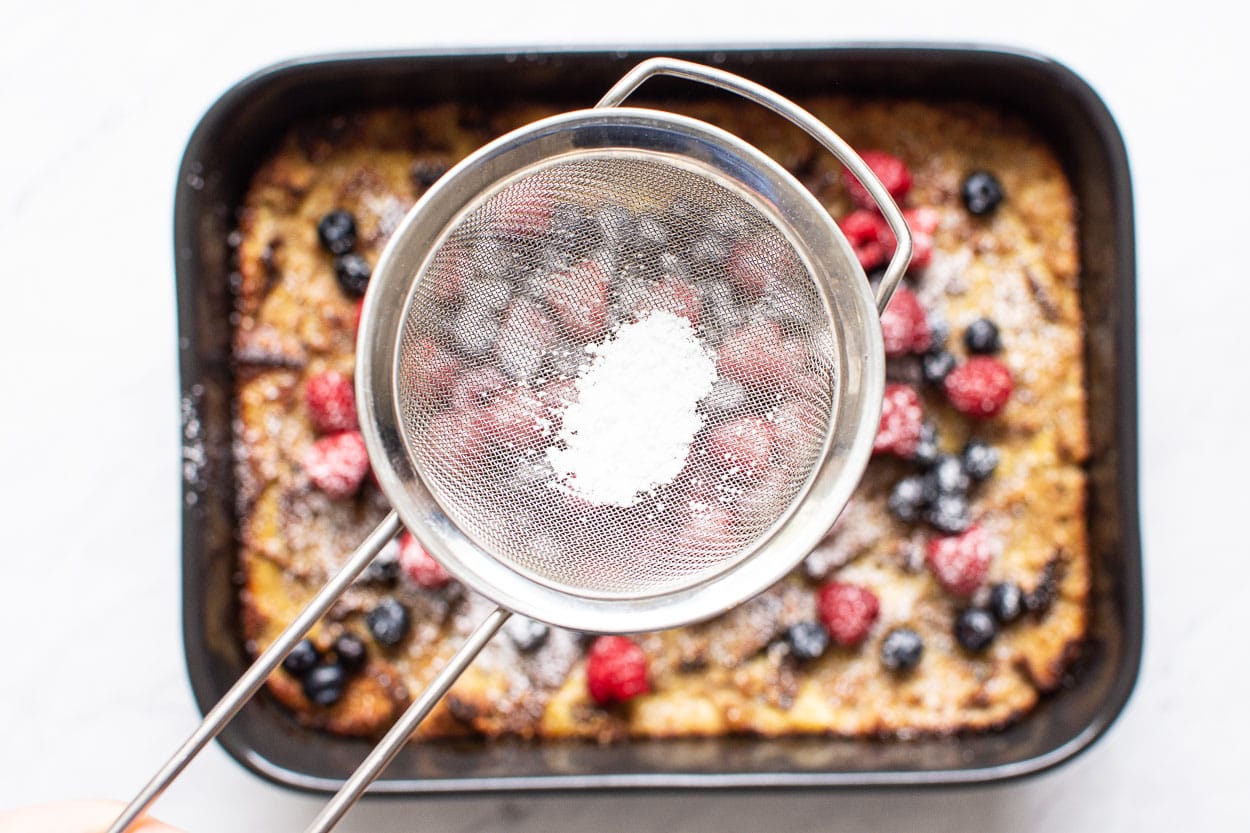 A mesh sifter with icing sugar over french toast casserole covered in berries.