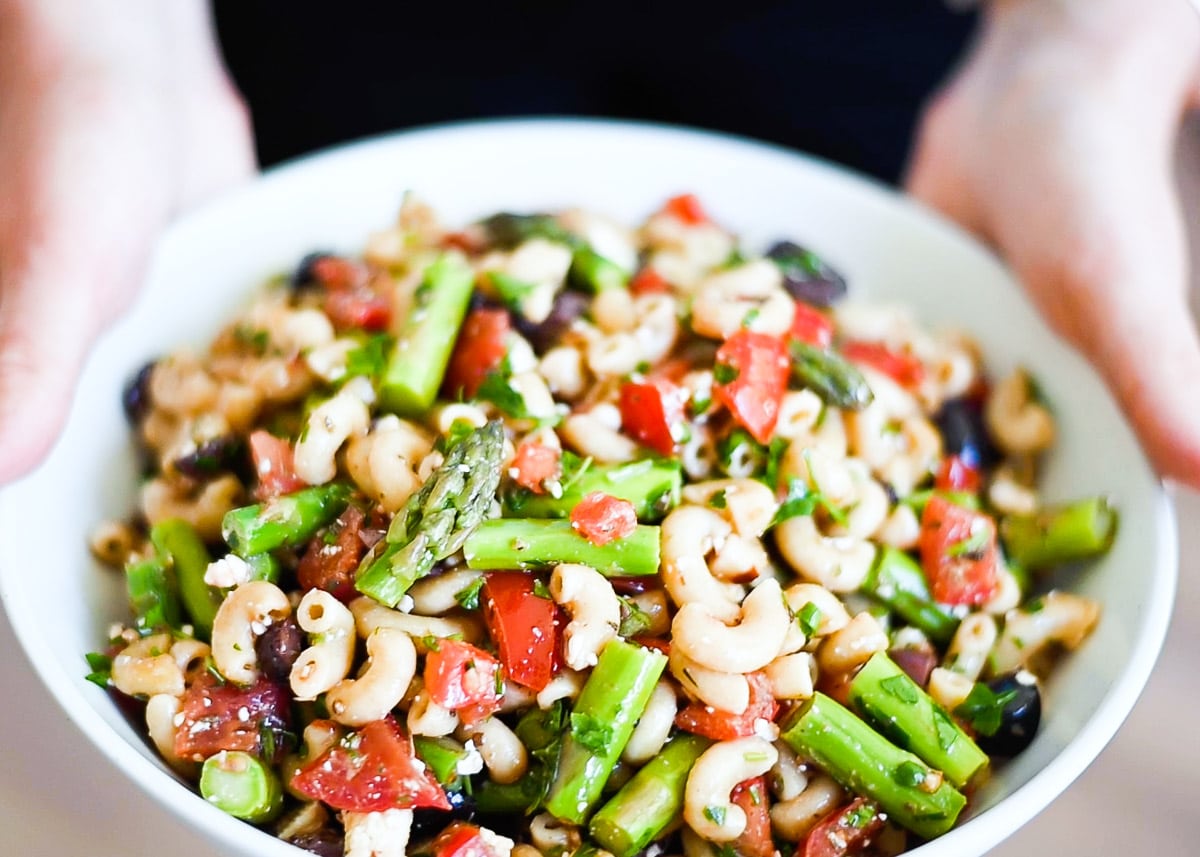 Hands holding healthy pasta salad in white bowl.