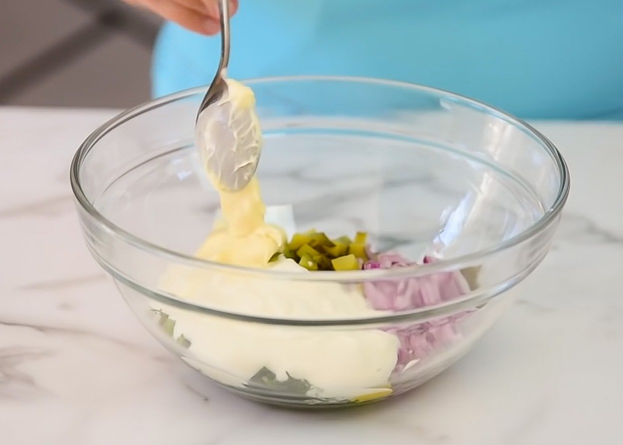 Adding mayo to a bowl with pickles, red onion, yogurt, dill and spices.