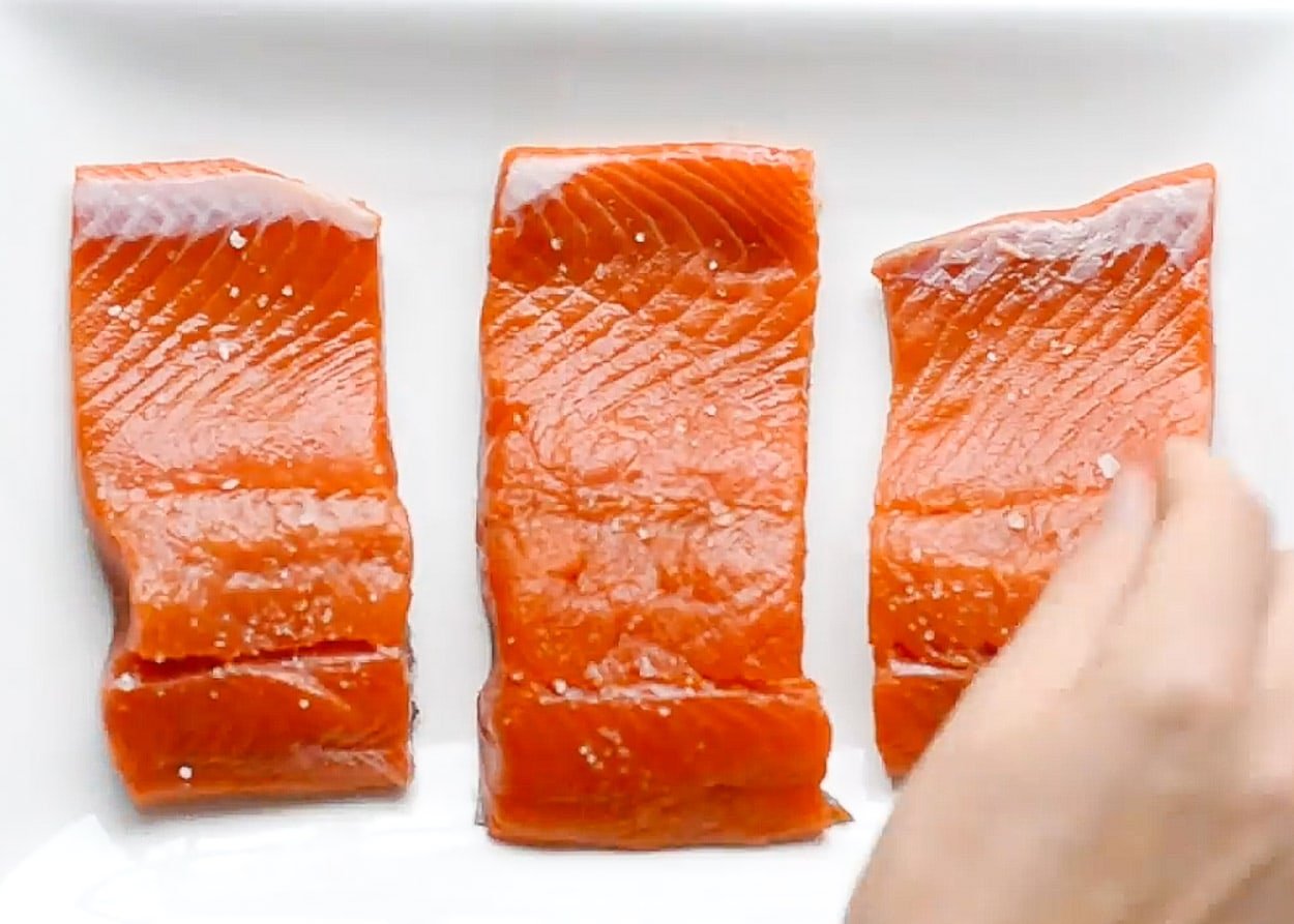 Salting three salmon fillets on white plate.