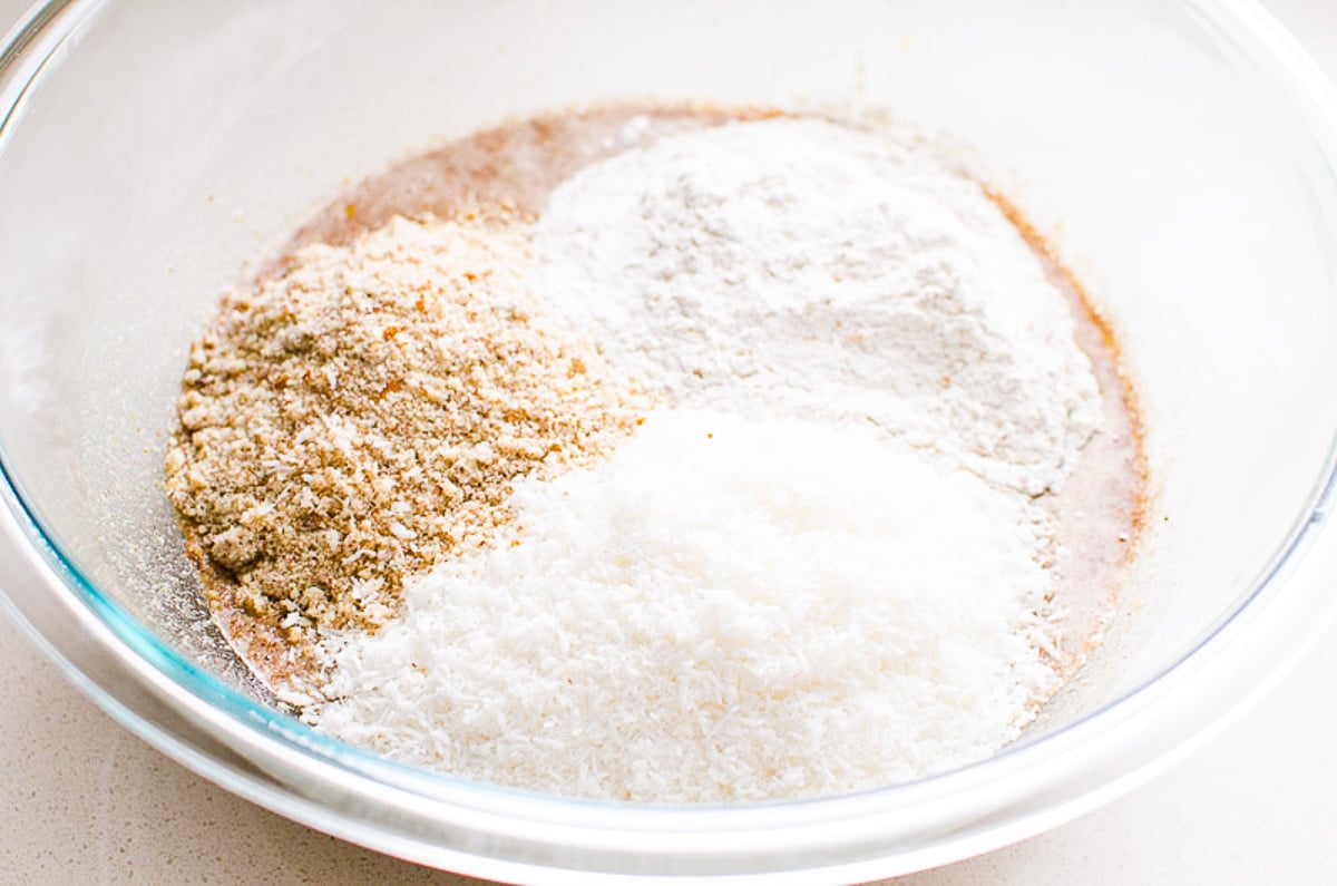 Adding coconut flakes, almond flour and whole wheat flour to a bowl with liquid ingredients.