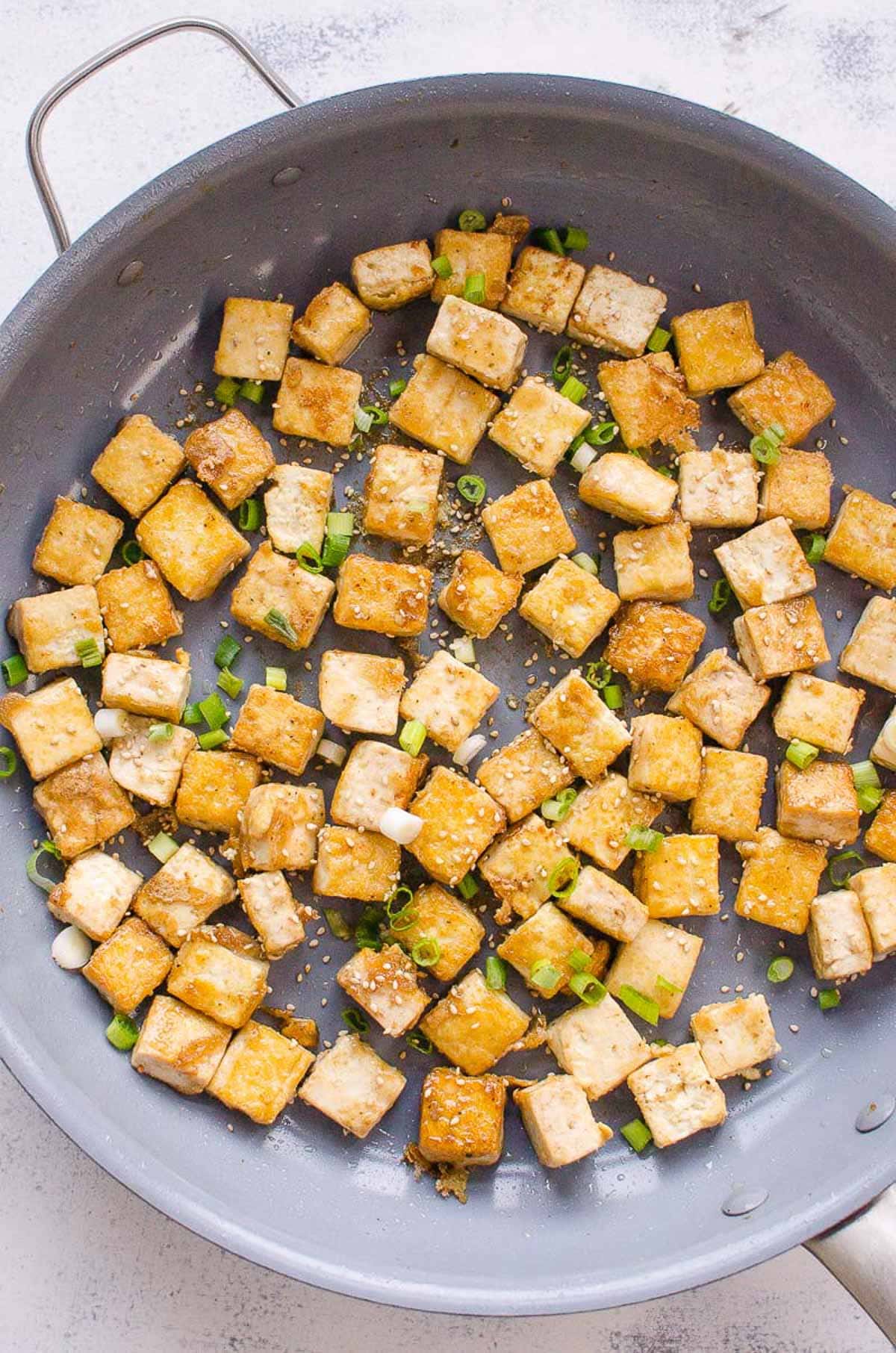 Crispy fried tofu on skillet garnished with green onions and sesame seeds.
