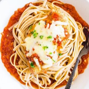 Healthy baked chicken parmesan served over a bed of spaghetti in white bowl with black fork.