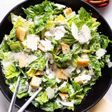 Healthy caesar salad with croutons and shaved parmesan cheese in black bowl with metal spoons.