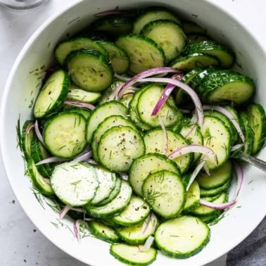 Healthy cucumber salad with red onions, dill, salt and pepper in white bowl with a spoon.