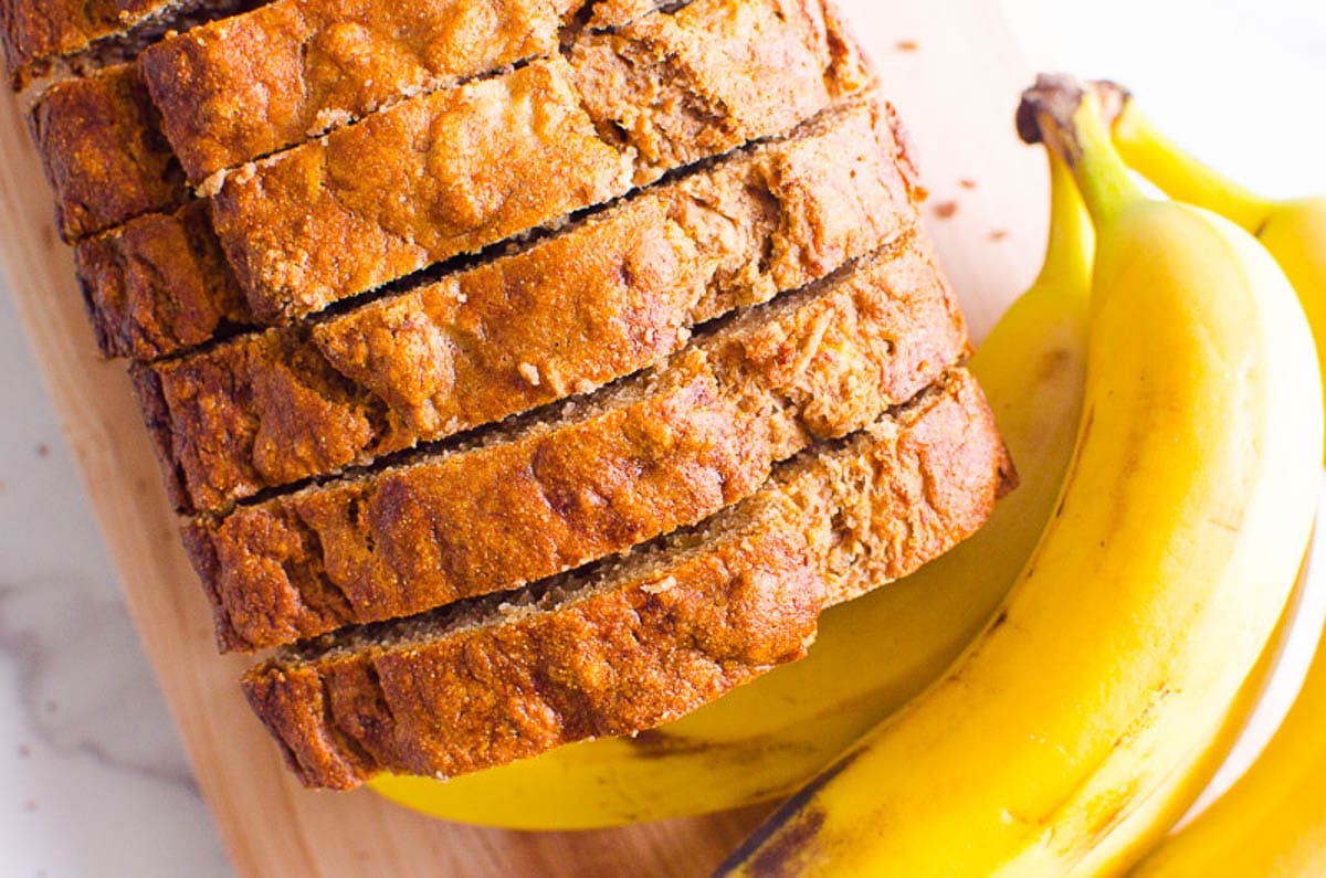 Healthy banana bread with applesauce sliced on a cutting board with three bananas.