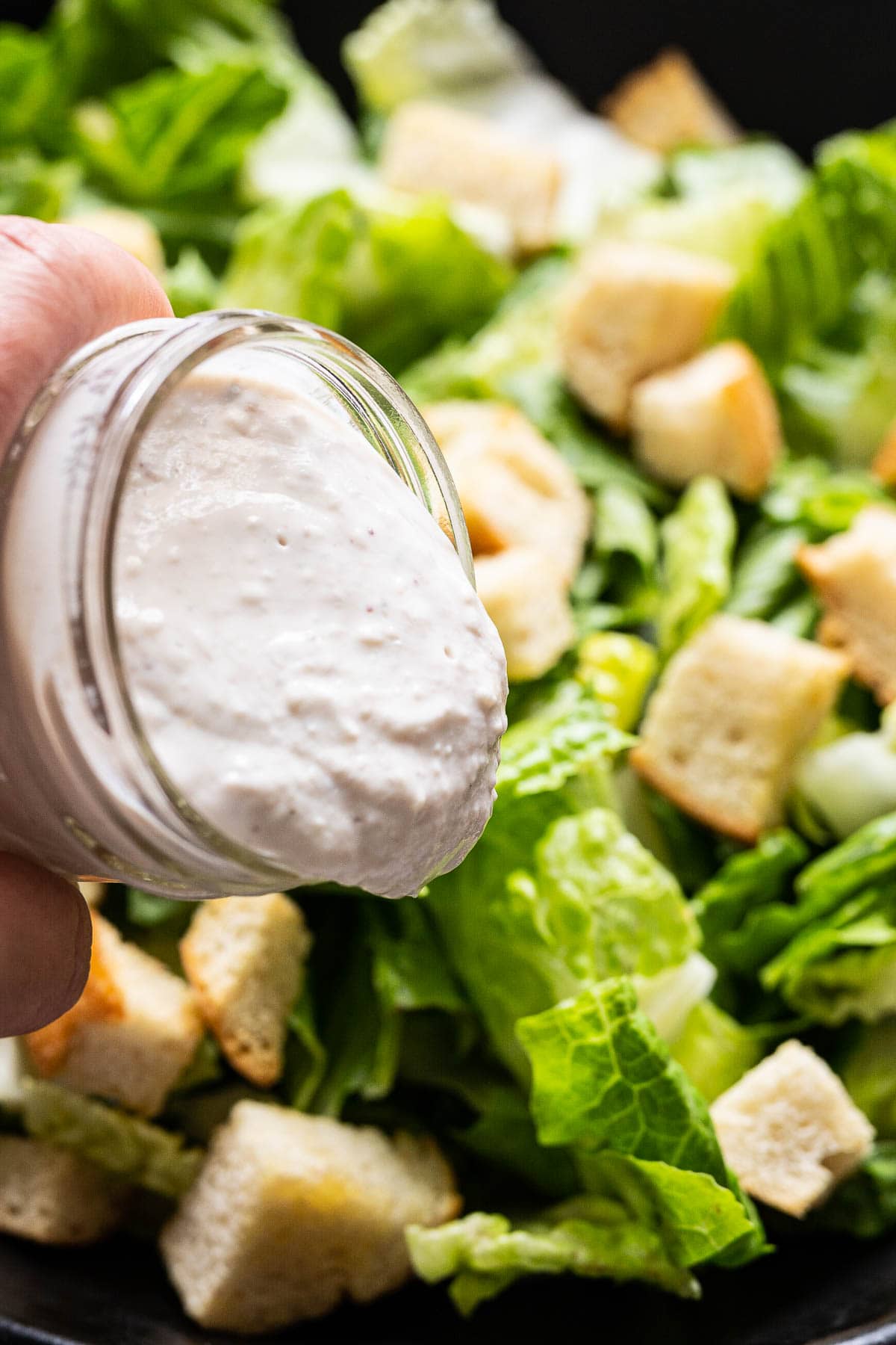 Pouring healthy caesar dressing over a bowl with Romaine lettuce, croutons and parmesan cheese.