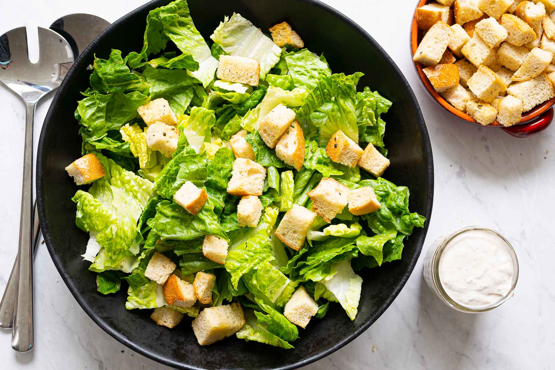 Romaine lettuce in bowl with croutons; metal spoons, caesar dressing in a jar, croutons in a bowl and on the counter.
