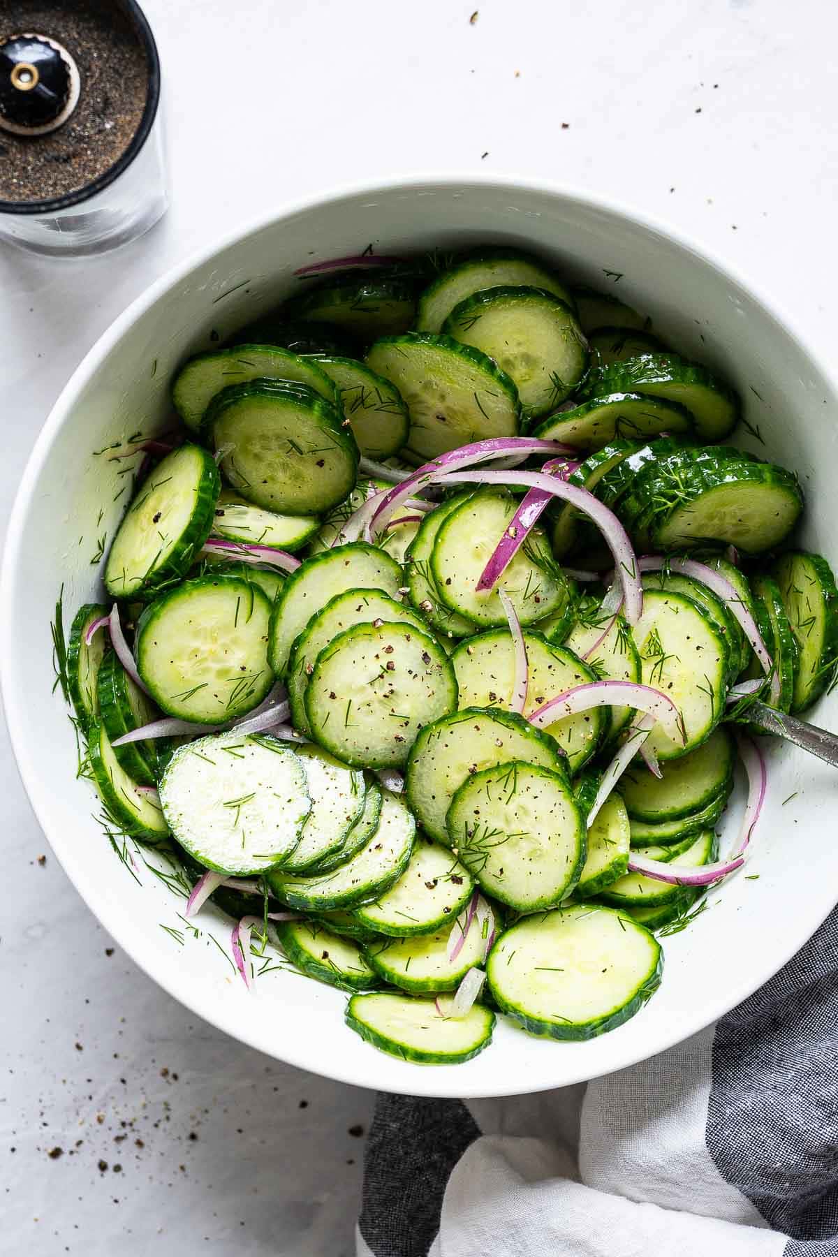 Healthy cucumber salad with red onions, dill, salt and pepper in white bowl with a spoon.