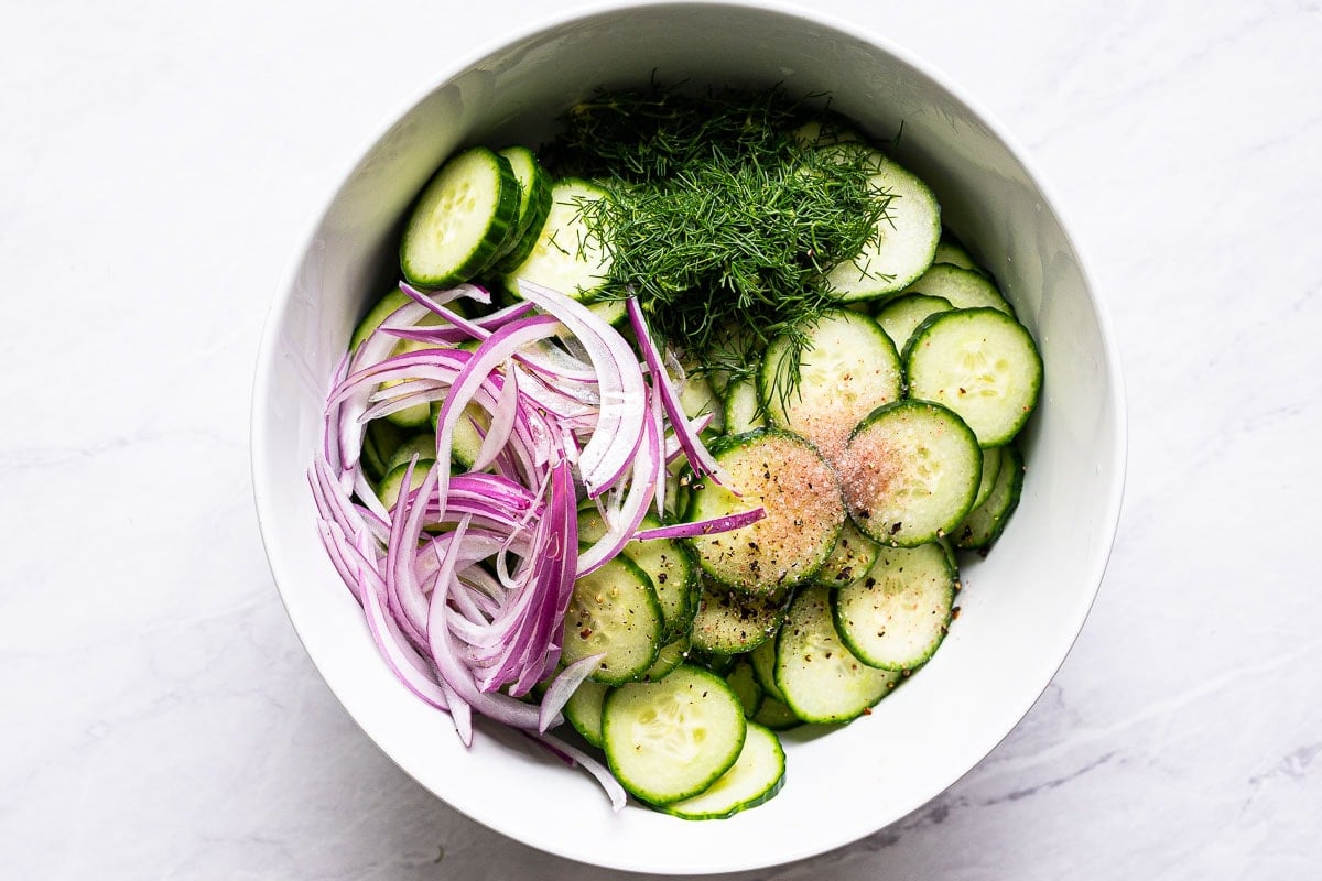 Sliced cucumbers and red onion, chopped dill, salt and pepper in white bowl.