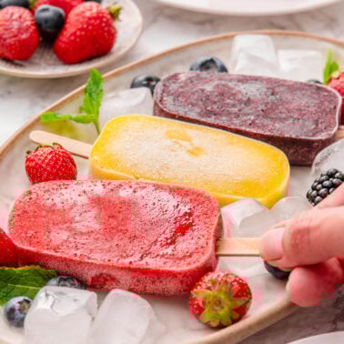 Three healthy popsicles on a platter with ice and berries. Person picking up one.