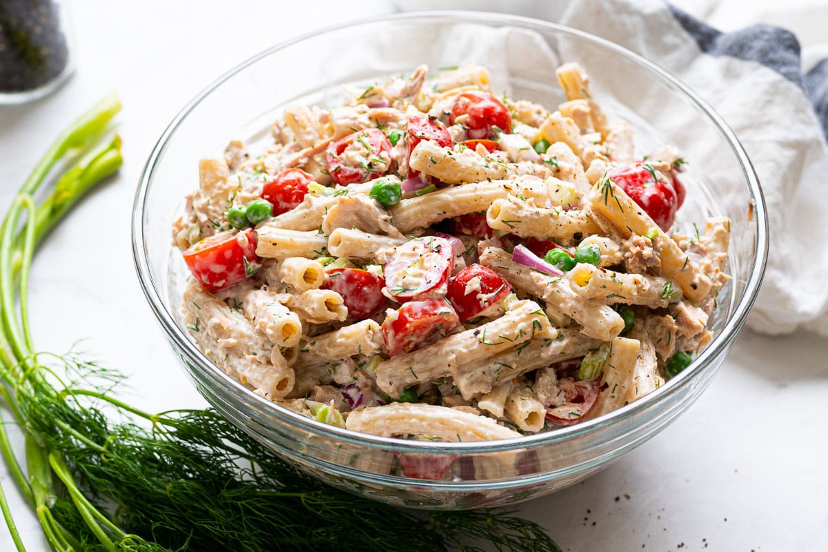 Pasta salad with tuna and yogurt dressing in a bowl with dill and a linen nearby on a counter.