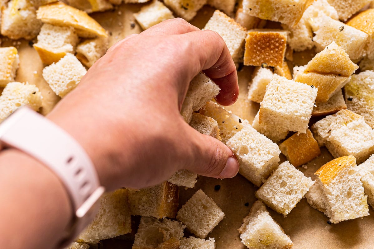 A hand mixing up seasoned bread cubes on baking sheet.