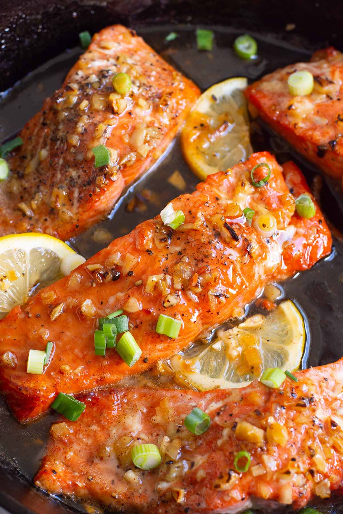 Honey garlic glazed salmon with green onion and lemon slices in cast iron skillet.