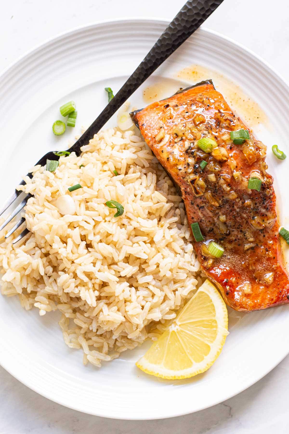 Honey garlic glazed salmon with brown rice and lemon wedge on white plate with black fork.