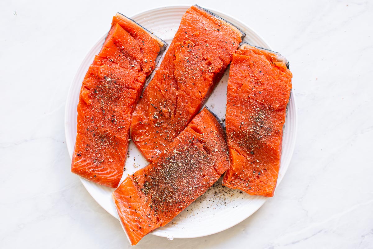 Four seasoned salmon slices seasoned with salt and pepper on white plate.